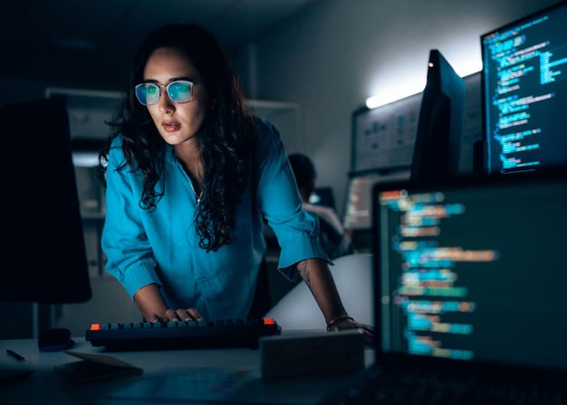 A female software engineer works on a project in the evening, and lines of code are visible on her monitors.