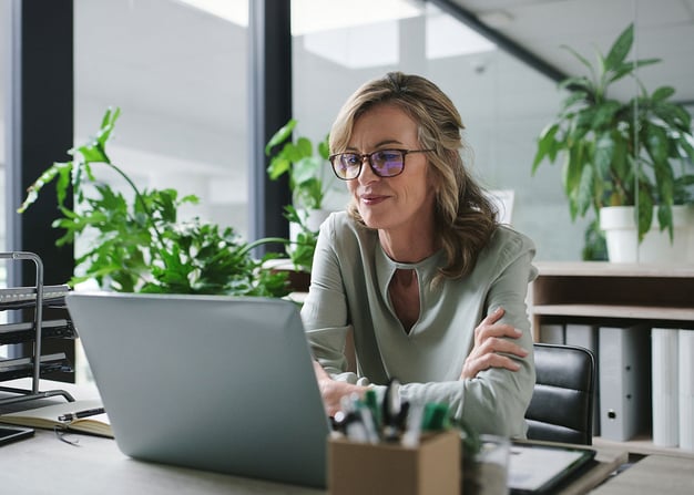 A woman working on a laptop in a well lit office with green plants.
