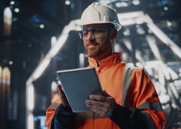 A manufacturing employee in a hardhat and orange jumpsuit uses a tablet.