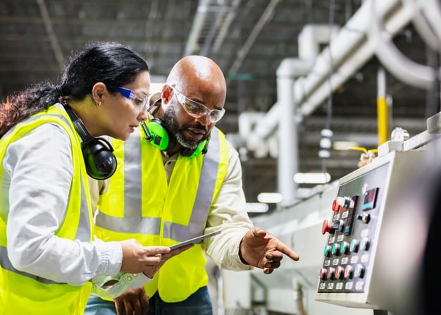 Two factory workers wearing yellow vests inspect equipment.